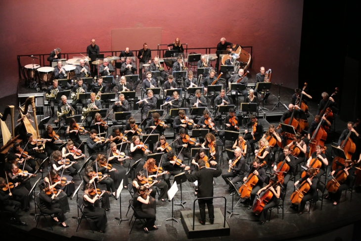 Un orchestre symphonique joue sur scène, dirigé par un chef d'orchestre vêtu de noir. Les musiciens, également en noir, sont répartis en sections : cordes, cuivres, bois, percussions et harpes. Le fond de la scène est rougeâtre.