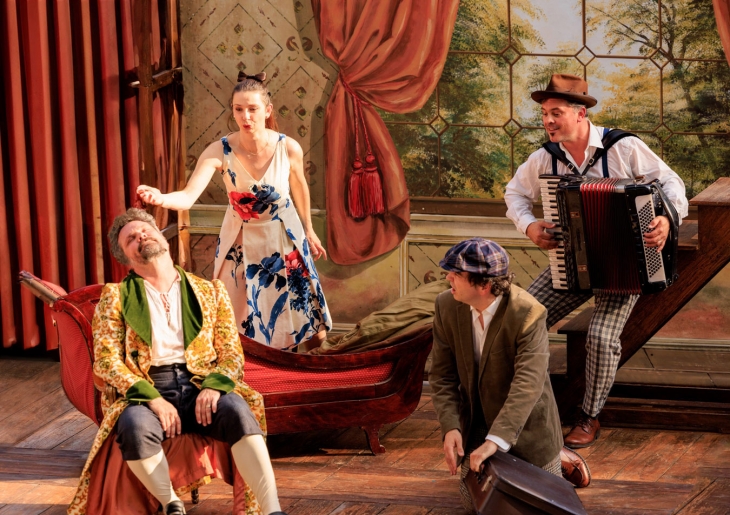 Une scène de théâtre avec quatre acteurs. Un homme en costume coloré est assis sur un canapé rouge, l'air exaspéré. Une femme en robe blanche à motifs floraux le réprimande, debout derrière lui. Un homme accroupi porte une valise marron et un chapeau à carreaux. À droite, un musicien en tenue rétro joue de l'accordéon.