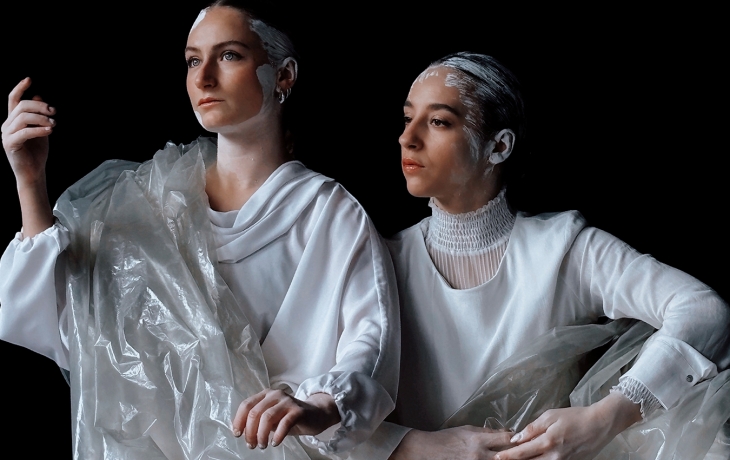 Deux interprètes sont sur scène, vêtues de blanc et enveloppées dans des voiles en plastiques blanc translucides. Elles ont les cheveux peints en blanc, comme du plâtre. elles sont de profil.