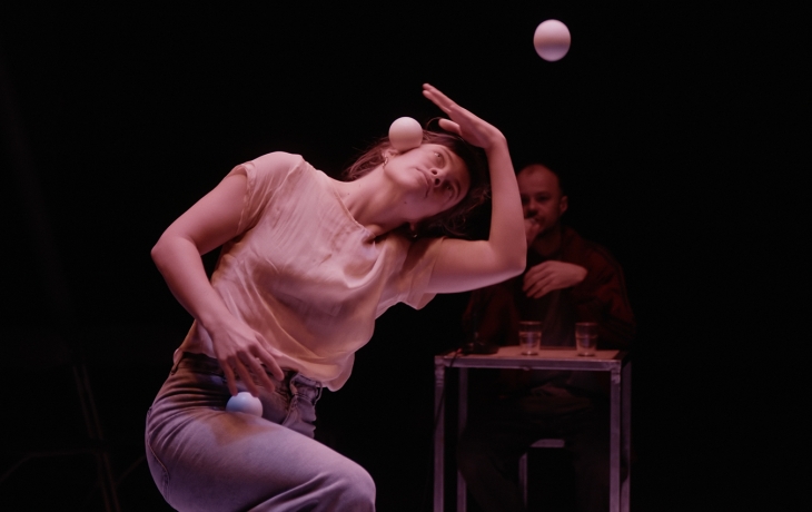 Suc scène, la jongleuse a la tête penchée et fait tenir en équilibre une balle. La deuxième balle est dans les airs et la troisième dans sa main. à l'arrière plan, en floue, il y a le rappeur à une table.