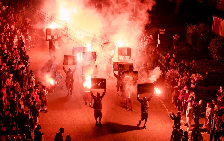 Dans la rue, le public laisse place a une déambulation chaotique. Les interprètes portent au-dessus de leurs têtes des tonneaux. Il y a beaucoup de fumée avec des fumigènes, les couleurs sont d'un rouge vif.