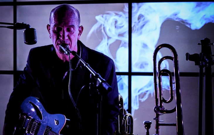 Un homme chante avec intensité dans un micro, en jouant de la guitare électrique, entouré de micros et d’un trombone posé à côté. Derrière lui, un grand écran lumineux projette des volutes de fumée bleutée, créant une ambiance mystérieuse et électrique.