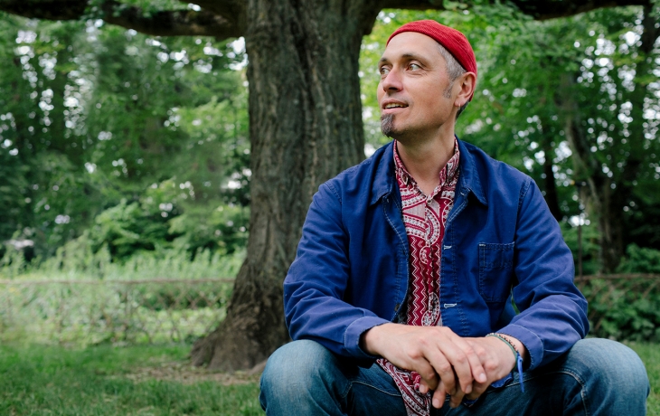 Dans la nature verdoyante, Thierry Combe est assis, au premier plan. Il porte une chemise bleu et un bonnet rouge. Il tourne la tête en rendant vers sa droite.