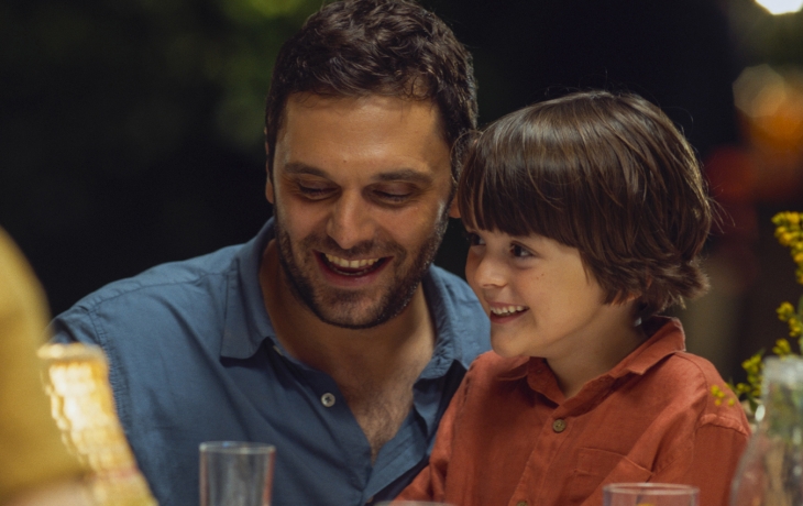 Un homme en chemise est à table, il porte un petit garçon sur ses genoux. Ils sourient.
