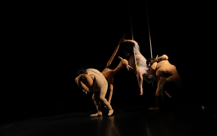 Sur scène, dans l'obscurité, deux interprètes en costumes couleurs chair sont en mouvement autour de la troisième interprète qui un costume en tulle blanc sur un agrès aérien.