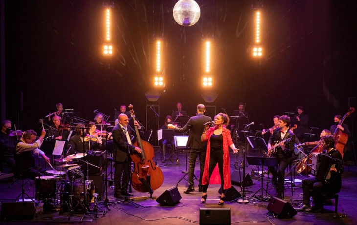 Sur une scène colorée, l'orchestre entoure la chanteuse qui est habillée en rouge vif. Au dessus d'eux se trouve une boule disco.