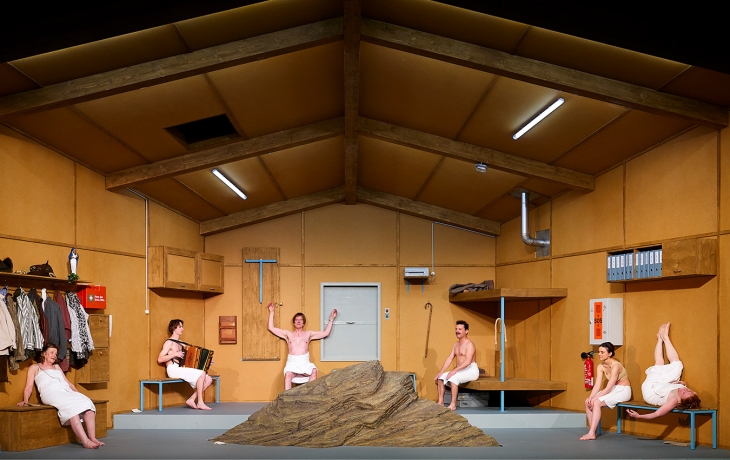 Photo de scène, dans un chalet, les sic interprètes sont tous éloignés les uns des autres, tous vêtues de serviettes blanches. Au centre, il y a un rocher.