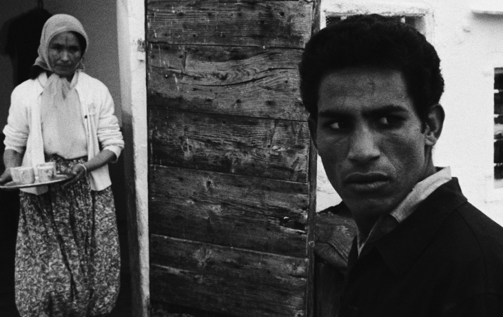 Photo en noir et blanc. Au premier plan à droite, un homme regarde sur la droite d'un air inquiet. À l'arrière plan, une femme marche en portant un plateau avec deux boissons.