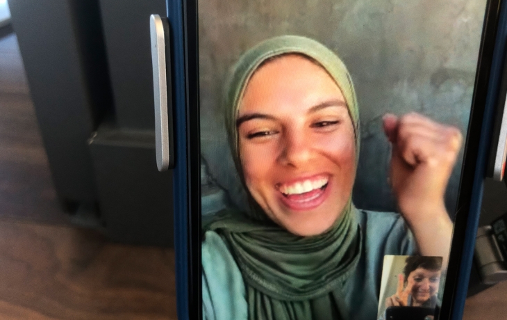 Un smartphone est posé sur un table, on voit la jeune photojournaliste Fatma Hassona en FaceTime avec Sepideh Farsi. Fatma sourit et lève le poing, déterminée. 