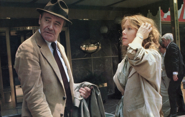 Un homme en costume et avec un chapeau est debout avec une femme blonde devant les portes d'un bâtiment. ils regardent derrière eux.