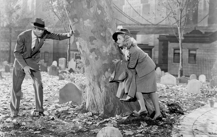 Photo en noir et blanc. Une femme se cache d'un homme derrière le tronc d'un arbre.