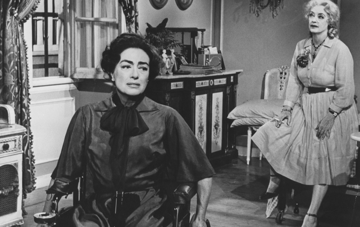 Noir et blanc. Au premier plan, Blanche est assise sur son fauteuil roulant et semble déboussolée. Derrière elle, Baby Jane fume une cigarette assises sur un lit, l'air pensante. 