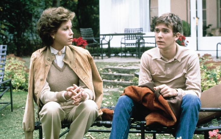 Le jeune garçon et sa mère sont assis sur un banc dehors. Elle le regarde tandis que lui regarde ailleurs.