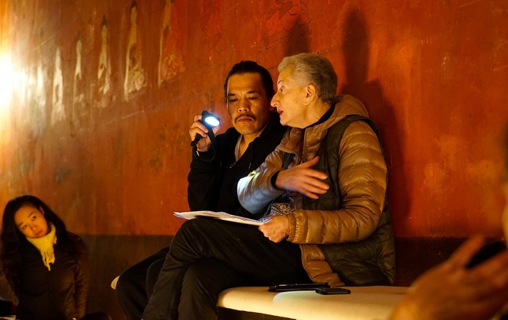 Assis contre un mur rouge, un homme et Hélène Cixous lisent des feuilles et discutent ensemble.