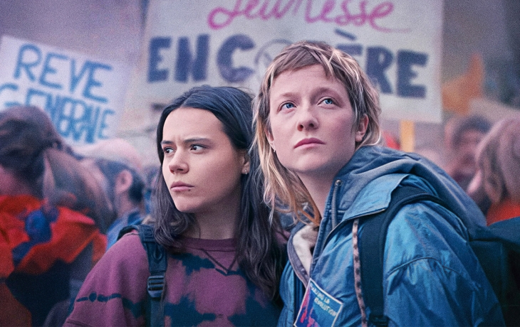 Deux jeunes femmes, une blonde et une brune, sont au centre d'une manifestation étudiante dans la rue. Au fond, on voit des pancartes dans la foule.