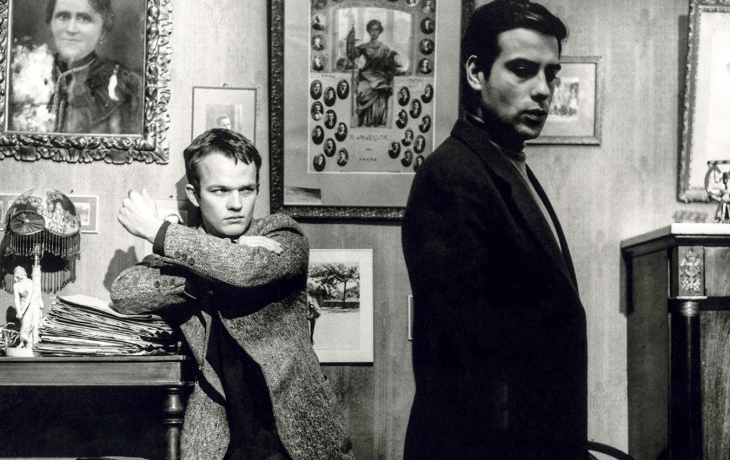 Noir et blanc. Le frère est assis à table et semble contrarié. Il a le poing serré.