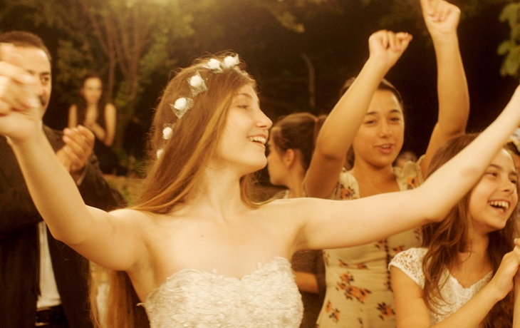 Gros plan sur une jeune fille qui danse les bras en l'air et qui porte une couronne de fleurs. Elle est entourée de ses soeurs.