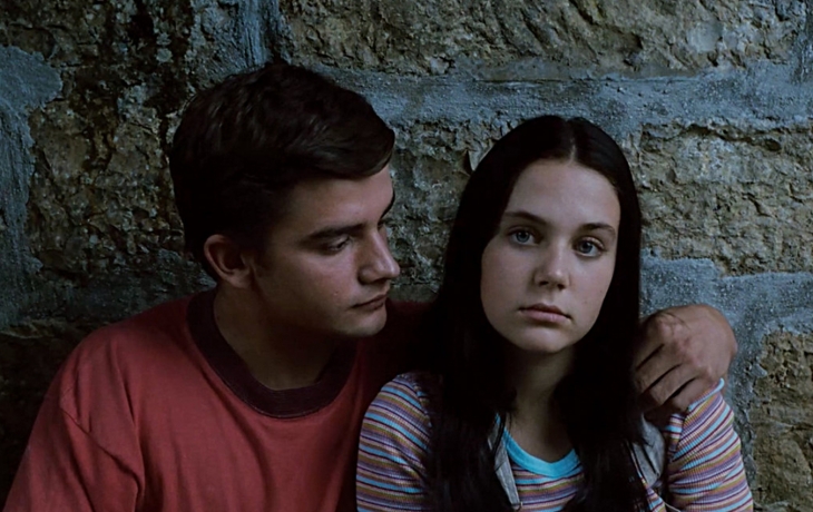 Contre un mur en pierre, un garçon et une fille sont côte à côte. Le garçon regarde la fille. La fille regarde l'objectif