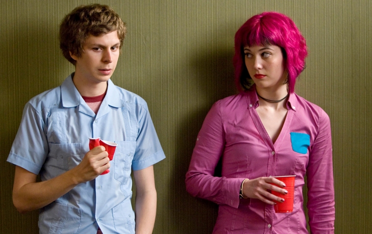 Contre un mur, un étudiant (Scott Pilgrim) est à côté d'une étudiante qui a les cheveux rose et une chemise rose. elle le regarde. Lui, serre son gobelet en plastique dans sa main.
