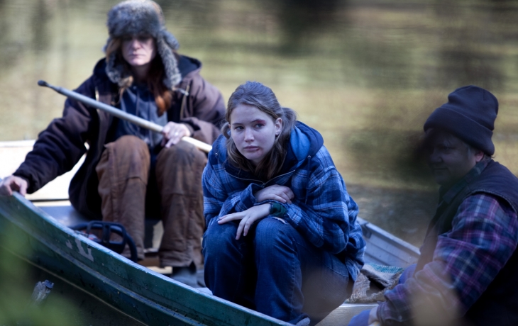 Sur une barque sur un lac, une jeune femme est recroquevillée, entourée de deux hommes qui mènent la barque près du rivage.