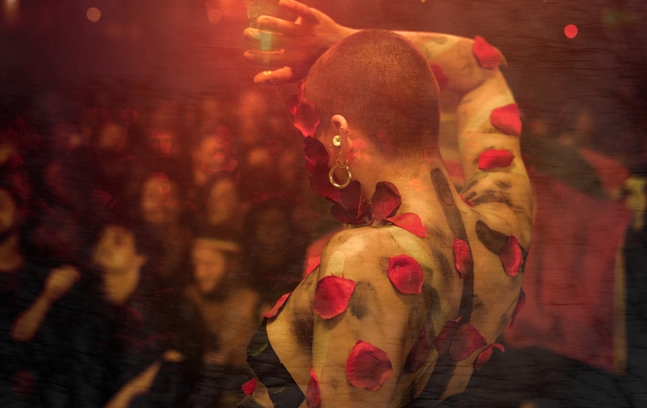 Vue de dos d’une personne sur scène, torse nu, la peau couverte de pétales rouges. Le bras est levé dans un geste de danse, avec un public flou et sombre en arrière-plan.