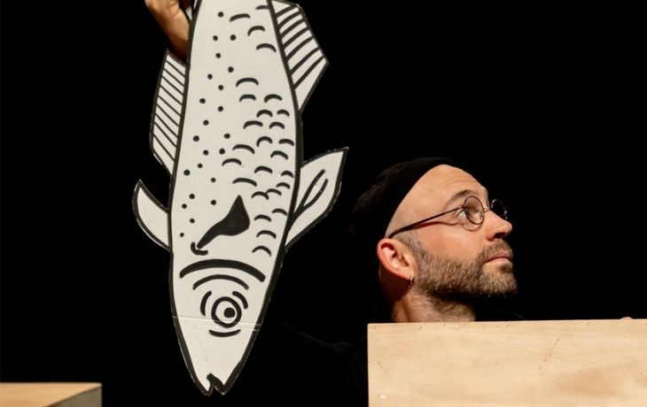 Un homme avec des lunettes se cache, une marionnette en forme de poisson devant lui 