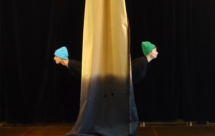Deux danseurs sont dods à dos derrière un tissus jaune qui descend du plafond 