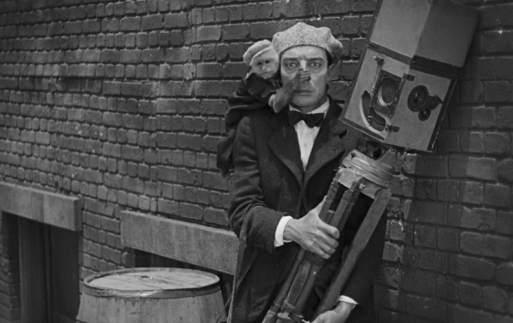Buster Keaton tient un trépied de caméra, un signe sur ses épaules lui recouvre le nez avec sa main 