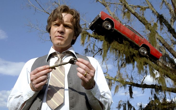 Un homme avec une clef, derrière lui, une voiture dans un arbre