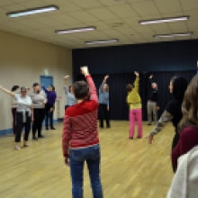 Un groupe d’adultes se tient dans une grande salle au sol en bois, disposés en cercle, et réalise un exercice corporel en levant un bras vers le haut.