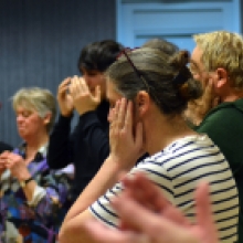 Plusieurs personnes sont alignées dans une salle et effectuent un exercice de concentration en posant leurs mains sur leurs oreilles ou leur visage. Les expressions sont calmes et attentives, suggérant un travail sensoriel.