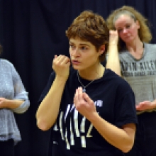 Trois femmes sont au premier plan dans une salle, chacune effectuant un geste près du visage ou du cou, comme dans un exercice guidé.