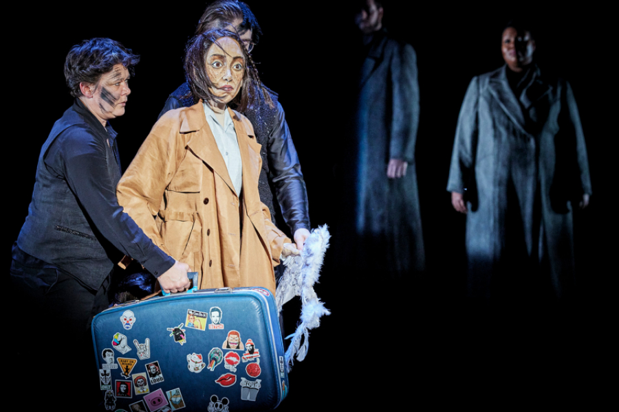 La marionnette au centre, éclairée, portant une valise. Une comédienne portant également cette valise. Au second plan, des comédiens dans l'ombre