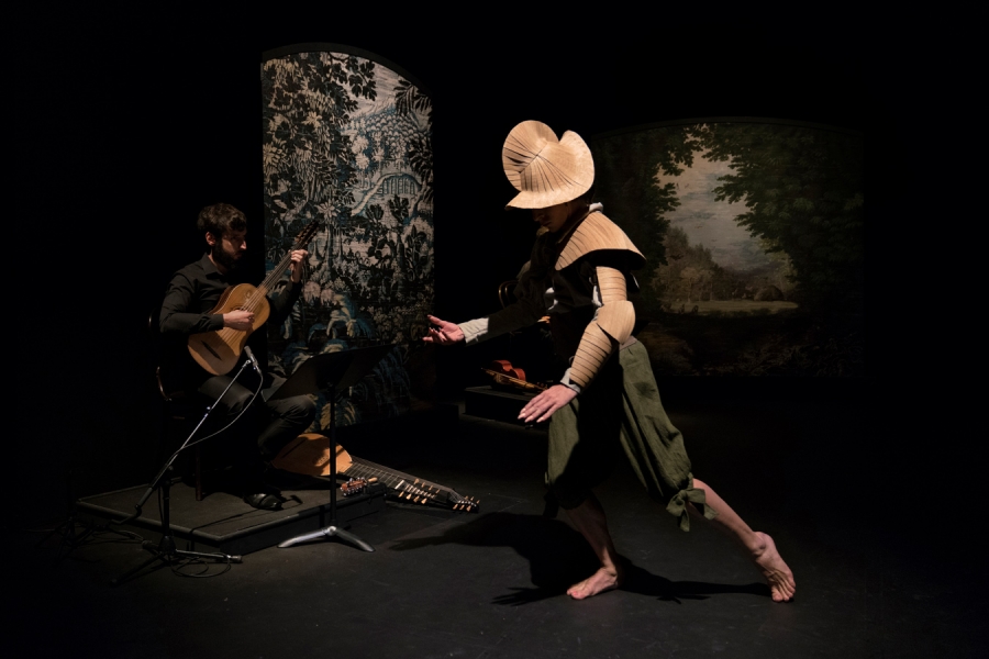  Un musicien en costume noir joue de la guitare classique assis à gauche de la scène. À droite, un danseur costumé de manière unique, portant un chapeau en forme de disque et des vêtements en papier plissé, exécute un mouvement de danse. Le fond est décoré de panneaux représentant des scènes naturelles.