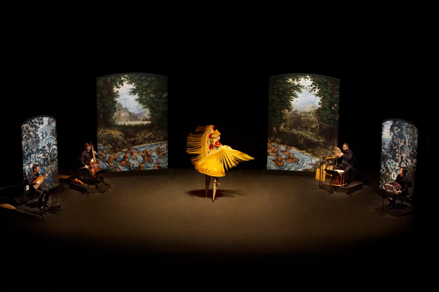 Une scène théâtrale sombre avec une danseuse au centre, portant un costume jaune vif à franges, effectuant une rotation. Quatre musiciens sont disposés autour de la scène, chacun jouant d'un instrument traditionnel. Le fond est composé de panneaux artistiques représentant des paysages naturels et des scènes mythologiques.