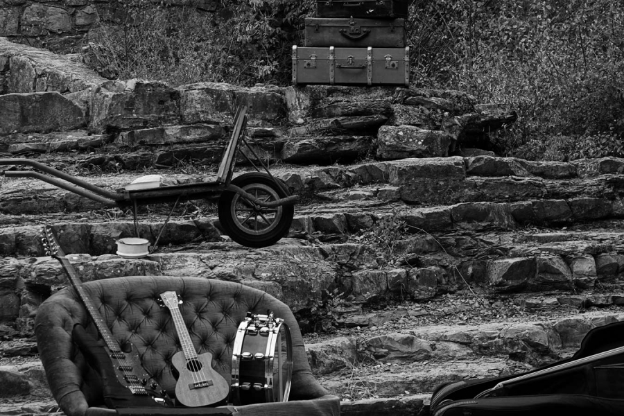 Photo en noir et blanc. On voit des escaliers en pierre dans la nature où il y a des instruments de musiques et des valises disposées un peu partout sur es marches.