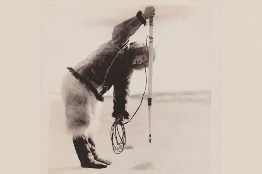 Photo en noir et blanc d'un esquimau qui pêche en faisant un trou dans la glace. Il est penché au dessus du trou et vise avec son harpon