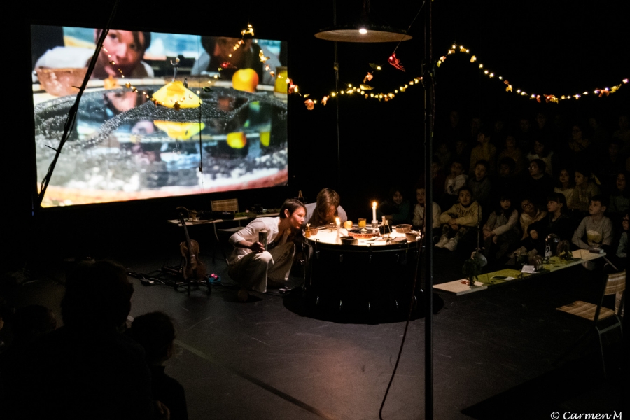 Deux personnes, penchées sur une table éclairée par des bougies, effectuent une performance devant un public assis. En arrière-plan, un grand écran projette une image agrandie d'une scène avec des fruits. Des guirlandes lumineuses décorent la scène.