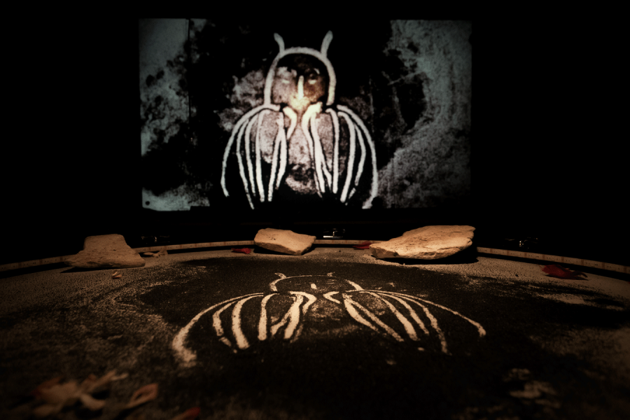 Un hibou stylisé est dessiné en sable sur une surface noire, avec une projection murale du même hibou en arrière-plan.