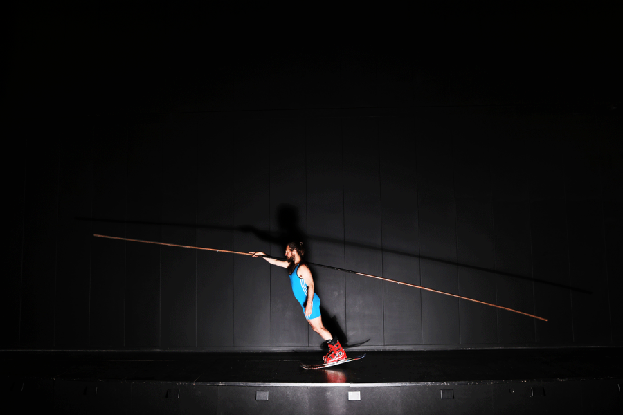 Un homme en combinaison de sport bleue et chaussures de ski rouges est en équilibre sur une planche, tenant une longue perche en bois. Il se trouve sur une scène de théâtre noire, éclairé par une lumière qui projette son ombre sur le mur derrière lui.