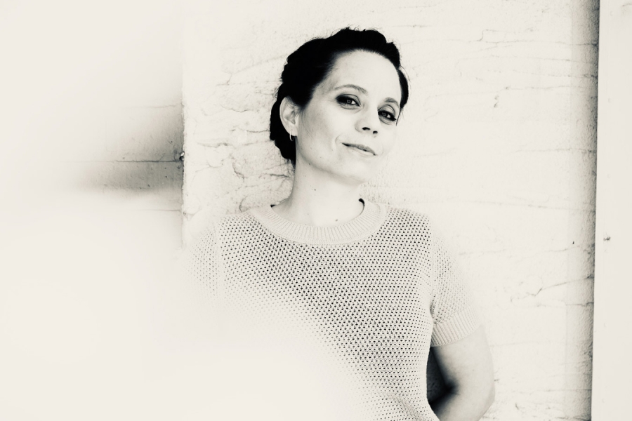 Portrait d'Anne Pacéo en noir et blanc. Elle est adossée à un mur blanc et regarde l'objectif en souriant