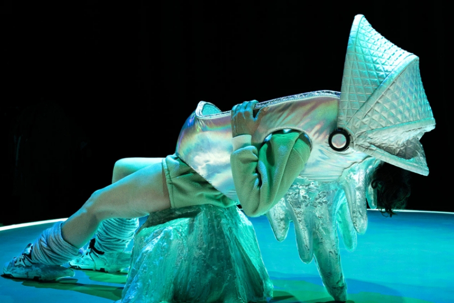 Un personnage incliné persque allongé dos au sol et retenu en équilibre par son costume avec des pics sur le dos. Les couleurs son bleutées et son costume métallique reflète cette lumière bleue