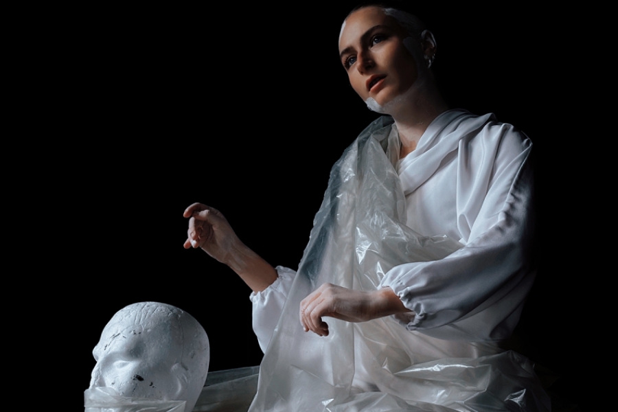 L'interprète est assise sur scène, ses bras en mouvement. Elle est vêtue d'habit blanc, enveloppée dans du papier plastique blanc translucide. Ses cheveux sont peint en blanc. Au sol à côté d'elle se trouve une sculpture d'un visage blanc.