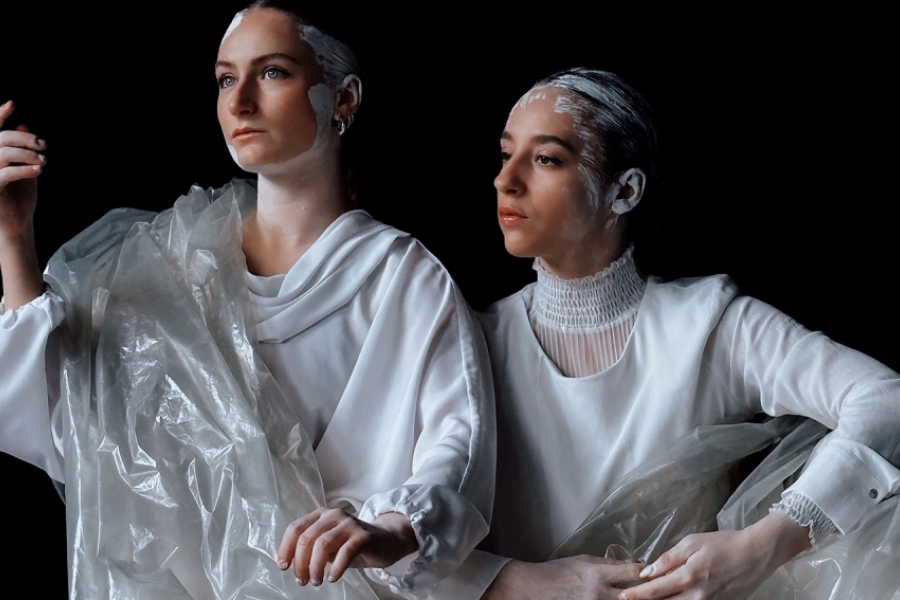 Deux interprètes sont sur scène, vêtues de blanc et enveloppées dans des voiles en plastiques blanc translucides. Elles ont les cheveux peints en blanc, comme du plâtre. elles sont de profil.