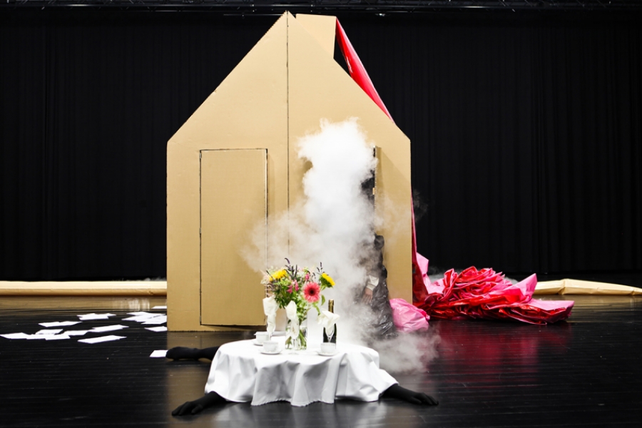 Une grande structure en forme de maison, faite de carton, se dresse sur scène. Devant elle, une table ronde blanche dressée avec un bouquet de fleurs semble posée sur un corps allongé au sol, tandis qu’un nuage de fumée s’échappe d’une porte entrouverte, créant un effet spectaculaire.