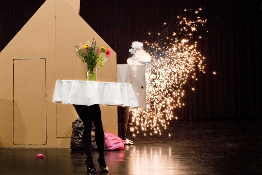 Une table ronde avec une nappe blanche et un bouquet de fleurs semble marcher sur des jambes humaines, tandis qu’une machine à effet déclenche une gerbe d’étincelles lumineuses à l’arrière-plan. La scène est étrange et joyeusement absurde, baignée d’un contraste entre quotidien et spectacle.