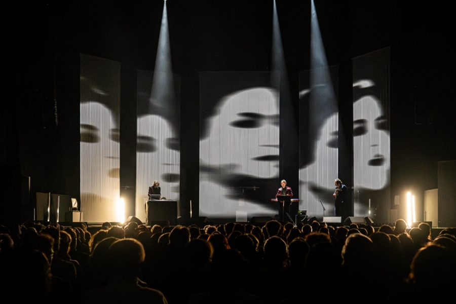 Photo de live, sur scène il y a les musiciens, au dessus d'eux est projeté un film en noir et blanc.