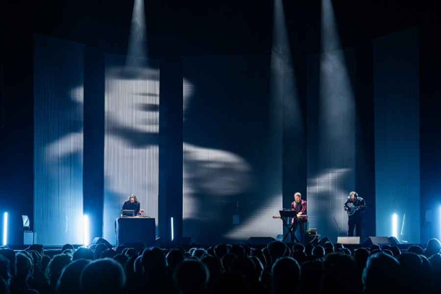Photo de live, sur scène il y a les musiciens, au dessus d'eux est projeté un film en noir et blanc.