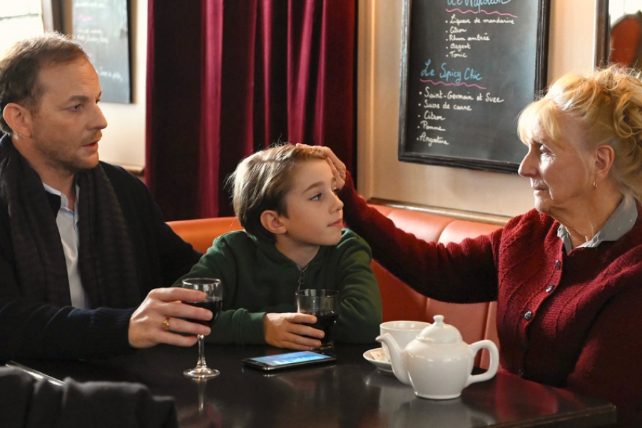 Dans un café, un homme, Michelle et son petit fils sont à table. Michelle fait un geste affectueux envers son petit fils. 