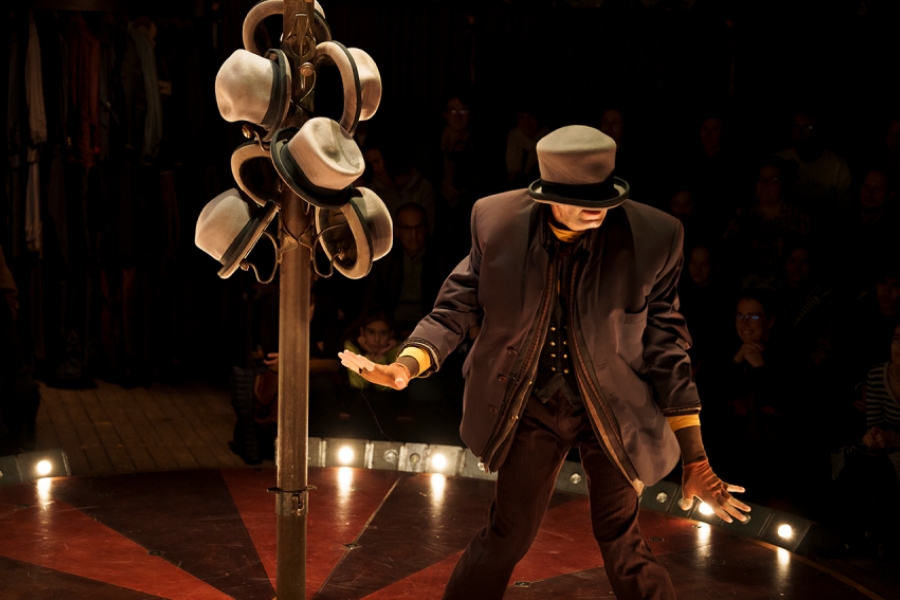 Dans un chapiteau, sur une petite scène circulaire, se trouve un porte-manteaux avec des chapeaux. À droite, se trouve un interprète en costumes et avec un chapeau enfoncé sur la tête, il ne voit rien et se déplace à l'aveugle.